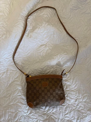Gucci brun axelväska med GG-mönster - Snygg brun axelväska från Gucci med klassiskt GG-monogram över hela väskan. Väskan har detaljer i ljusbrunt läder, justerbar axelrem och dragkedja upptill. Perfekt för dig som gillar tidlös design och vill ha en ikonisk accessoar. Obs ÄKTA vintage från 80/90 talet. , skriv om du har mer frågor 😁