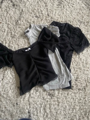 Tre trendiga toppar i svart och grått - Säljer tre snygga toppar: en svart plisserad topp med puffärmar, en grå enkel t-shirt och en svart spetsdetaljerad topp. Perfekta för att mixa och matcha till olika outfits. Alla har kort ärm och modern passform.