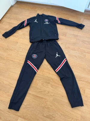 PSG x Jordan marinblå träningsset - Snyggt svart träningsset från PSG x Jordan med jacka och byxor. Setet har vita och röda ränder på ärmar och ben, PSG logga. Setet är inte äkta. Perfekt för fotbollsträning eller chill. Dragkedja på jackan och resår i midjan på byxorna. Storlek 164 men använde det när jag var 9 då jag var 137cm ungefär! Tar bara betalningar genom swish!