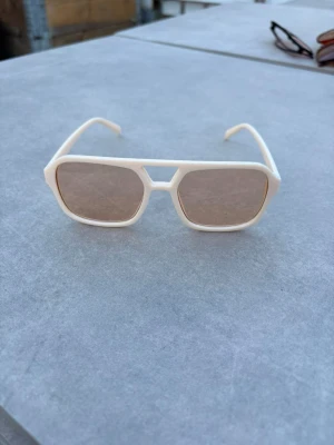 Vita retro solglasögon med brun ton - Snygga solglasögon med vit, kantig båge och bruna tonade glas. Modellen har en cool retrostil med breda skalmar och markerad näsbrygga. Perfekta för dig som vill sticka ut med en unik accessoar i sommar.