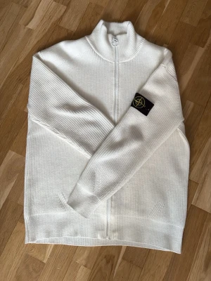 Vit stickad ziptröja från Stone Island - Vit stickad tröja från Stone Island med hel dragkedja och hög krage. Tröjan har ribbad struktur, två fickor framtill och den klassiska svarta logotypen på ärmen. Perfekt för lager-på-lager och snygg till jeans eller chinos.