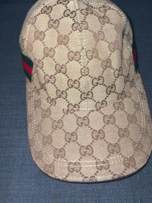 Beige keps från Gucci med GG-mönster - Snygg beige keps från Gucci med klassiskt GG-monogram över hela kepsen. På sidan finns den ikoniska gröna och röda randen. Kepsen har böjd skärm och är tillverkad i canvasmaterial. Perfekt för dig som vill sticka ut med en lyxig accessoar.