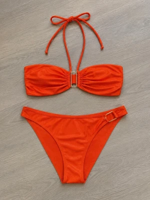 Gian Bandeau bikini med gulddetaljer - Snygg röd/korall/orange bikini med bandeau-topp och knyt i nacken. Toppen har guldfärgad detalj framtill och bikinitrosan har matchande guldfärgad detalj på sidan. Båda delarna säljs tillsammans och är i strl M! Den har endast blivit testad, annars inte använd!✨
