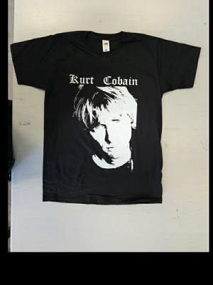 Svart Kurt Cobain t-shirt Fruit of the Loom - Svart t-shirt med stort vitt tryck av Kurt Cobain på framsidan och text i gotisk stil. Klassisk rund hals och korta ärmar. Tillverkad i mjuk bomull som känns skön mot huden. Perfekt för dig som gillar bandmerch och vill ha en ikonisk look. Helt oanvänd t-shirt med tryck Storlek: Smal 100% bomull Screentryckt