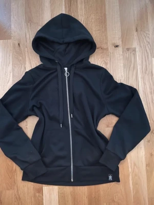 Svart hoodie med dragkedja FB Sister - Klassisk svart hoodie från FB Sister i storlek XS. Hoodien har dragkedja framtill, justerbar huva med snören och en stor ficka på magen. Tillverkad i mjukt material som passar perfekt till en avslappnad stil.