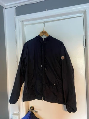 Mörkblå vindjacka från Moncler - Snygg mörkblå vindjacka från Moncler med huva och dragkedja framtill. Jackan har Moncler-logga på ärmen, justerbar huva med rödvit detalj och två fickor med dragkedja. Perfekt för blåsiga dagar och enkel att matcha till olika outfits. storlek 2 lika med m