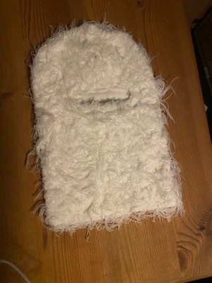 Vit fluffig balaclava / skidmask - Säljer en vit, superfluffig mössa med lång lugg och unik textur. Mössan har en cool, busig look tack vare de långa, mjuka fibrerna som ger en extra dimension. Perfekt för dig som vill sticka ut och hålla dig varm under kalla dagar. Aldrig använd - Helt oanvänd 