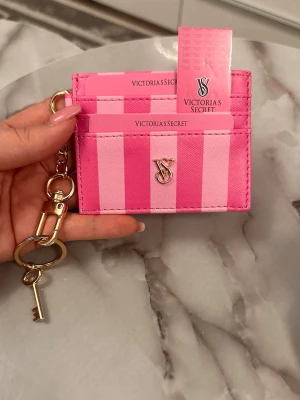 Rosa kortfodral från Victoria's Secret - Snyggt randigt kortfodral i rosa nyanser från Victoria's Secret. Fodralet har flera kortfack, ett metalliskt VS-emblem framtill och en praktisk nyckelring med karbinhake. Perfekt att ha i väskan eller hänga på nyckelknippan.