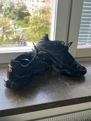 Nike Air Max Plus TN svarta sneakers - Säljer ett par svarta Nike Air Max Plus TN sneakers med klassisk TN-logga på hälen. Skorna har en sportig design med synliga luftkuddar i sulan och detaljerade sömmar. Materialet är en mix av syntet och mesh för bra andningsförmåga och komfort.