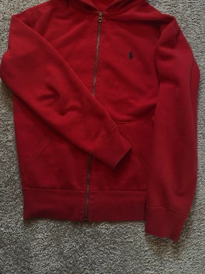 Röd hoodie från Polo Ralph Lauren - Snygg röd hoodie från Polo Ralph Lauren med dragkedja och klassisk blå logga broderad på bröstet. Hoodien har huva, fickor framtill och ribbade muddar vid ärmslut och nederkant. Perfekt för en avslappnad och stilren look.