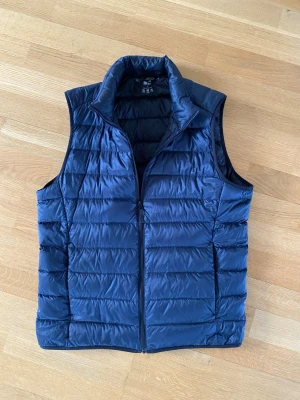 Mörkblå dunväst från Uniqlo strl S - Snygg och lätt mörkblå dunväst från Uniqlo med hög krage och dragkedja framtill. Västen är quiltad och har en stilren, minimalistisk design. Perfekt att slänga över en hoodie eller tröja för extra värme. Materialet är tunt men isolerande. Fint skick utan några defekter dessutom. Hör av er vid frågor och funderingar. 
