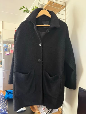 Svart ullkappa från Monki - Stilren svart kappa från Monki med två stora fickor framtill och klassisk krage. Kappan har en rak, oversized passform och stängs med knappar. Materialet är ullblandning. Känns mer som en M än S i storleken, eftersom att den är oversize