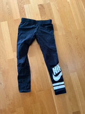 Svarta leggings från Nike med tryck - Svarta leggings från Nike med vitt logotryck och två vita ränder på ena benet. Mjukt och stretchigt material som sitter skönt. Perfekta för en sportig look eller till chill-outfiten. Resår i midjan för extra komfort.