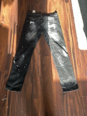 Svarta dsquared2 jeans - Säljer ett par svarta  jeans med coola slitningar och vita färgstänk. Jeansen har klassisk femficksdesign och smal passform. Perfekta för dig som gillar en edgy och trendig streetstil.
