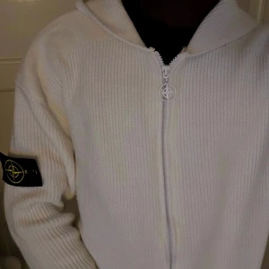 Vit hoodie med dragkedja från Stone Island - Vit ribbstickad hoodie från Stone Island med dragkedja framtill och klassisk logotyp-patch på vänster ärm. Tröjan har huva och är tillverkad i ett mjukt, stickat material som ger en clean och stilren look.