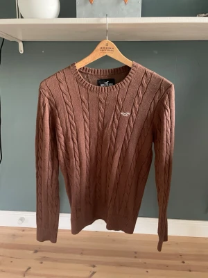 Brun kabelstickad tröja Hollister - Säljer en brun kabelstickad tröja från Hollister med rund halsringning och diskret logga broderad på bröstet. Tröjan har långa ärmar och ribbade muddar vid ärmslut och nederkant. Perfekt för dig som gillar klassisk och enkel stil. Endast testad