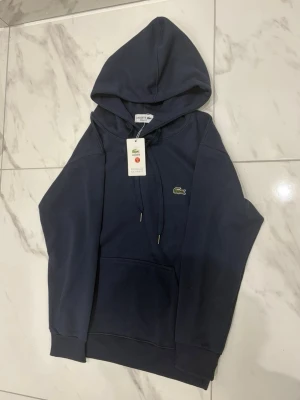 Mörkblå hoodie från Lacoste - Snygg mörkblå hoodie från Lacoste med klassisk krokodillogga på bröstet. Hoodien har dragsko i huvan och en stor magficka framtill. Tillverkad i mjukt material som känns skönt mot huden. Perfekt för en avslappnad och stilren look.