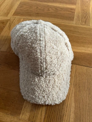 Beige teddykeps - Mysig beige keps i teddyfleece med böjd skärm och justerbar kardborreband bak. Kepsen har fluffig modern textil  som sticker ut och passar till många outfits.