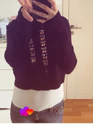 Svart cropped hoodie med snörning - Säljer en svart cropped hoodie med cool snörning framtill och breda muddar. Hoodien har en klassisk huva och är gjord i mjukt stickat material. Perfekt för en avslappnad och trendig look.Har tagit av snöret men skickar med det i paketet