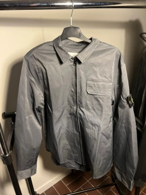 G Stone Island - Snygg grå overshirt från Stone Island med klassisk krage, bröstficka och dragkedja framtill. Långärmad modell med ikonisk logotyp på ärmen. Perfekt för lager-på-lager och streetwear-stil.