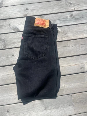 Svarta Levi'S 501 - Säljer ett par svarta jeans från Levi's med klassisk rak passform. Jeansen har fem fickor, Levi's ikoniska läderpatch bak i midjan och röd logotagg på bakfickan. Perfekta för en stilren och avslappnad look.