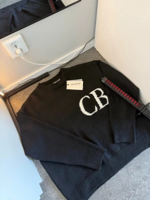 Svart stickad tröja Cole Buxton - Svart stickad tröja från Cole Buxton med stora vita bokstäver 'CB' på bröstet. Tröjan har rund halsringning och ribbstickade muddar vid ärmslut och nederkant. Perfekt för en stilren och trendig look. Perfekt för alla årstider. Priset går absolut att diskuteras