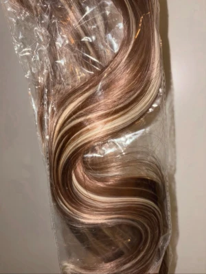 Brun-blond hårförlängning med clips 2st - Snygg hårförlängning med clips i en mix av bruna och blonda slingor. Långt, vågigt syntethår som ger ett naturligt intryck och extra volym. Perfekt för att snabbt förändra din look. Levereras i förpackning.