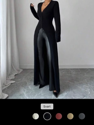 Svart lång jumpsuit med v-ringning - Trendig svart jumpsuit med lång, figurnära överdel och vida ben. Djup v-ringning framtill och knäppning längs överdelen. Materialet ser ut att vara en mix av stretchigt tyg och eventuellt konstläder på benen. Perfekt för dig som gillar en stilren och edgy look. Är helt ny skicka gärna pris förslag!!