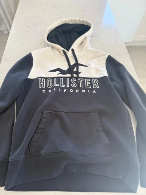 Blå och vit hoodie från Hollister - Snygg hoodie från Hollister i blå och vitt med stor logga och texten 'Hollister California' på bröstet. Hoodien har huva med svarta snören och en stor magficka. Perfekt för en avslappnad och trendig stil.
