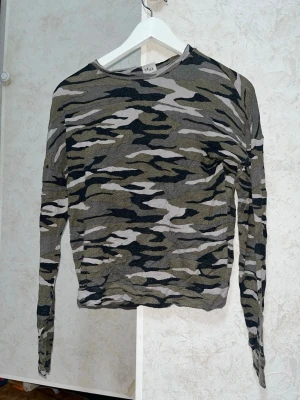 Camoflauge färgad långärmad tröja Gina Tricot - Säljer en långärmad tröja från Gina Tricot i camoflaugemönster med nyanser av grått, svart och grönt. Modellen är figurnära med rund halsringning och mjukt, stretchigt material. Perfekt för dig som gillar streetstyle och vill sticka ut med ett coolt mönster.