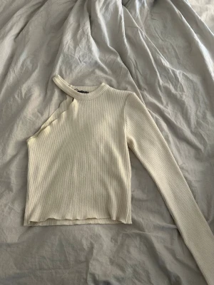 Off shoulder topp Zara - Trendig ribbad topp i beige med en ärm och offshoulder-design. Croppad modell med smal passform och rund halsringning. Perfekt för dig som gillar unika detaljer och stilren look.