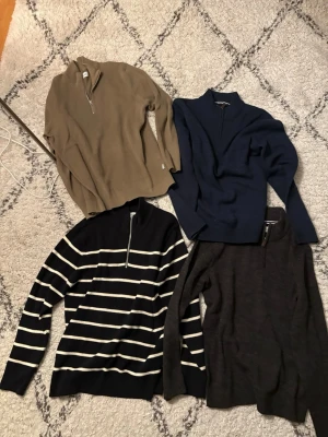 Fyra stickade tröjor med dragkedja - Fyra stickade tröjor (herr) med halv dragkedja i olika färger: beige, marinblå, mörkgrå och en svart med vita ränder. Alla har lång ärm och klassisk krage. Perfekta för lager på lager och stilrena outfits. Storlek XL. Säljer för 175kr/st. Mycket bra skick