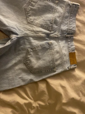 Ljusblå jeans från Gina Tricot - Säljer ett par ljusblå jeans från Gina Tricot med raka ben. Jeansen är baggy med slitningar och hål. Jeansen har klassiska fem fickor, bälteshällor och knappgylf. Perfekta för en avslappnad och trendig look.