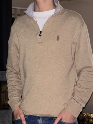 Beige halfzip från Polo Ralph Lauren - Snygg beige halfzip tröja från Polo Ralph Lauren med halv dragkedja och broderad logga på bröstet. Tröjan har ribbade muddar och krage med grå insida. Perfekt till jeans för en clean och stilren look. 
