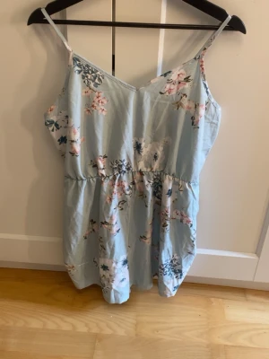 Ljusblå blommig somrig byxdress - Söt ljusblå byxdress med smala axelband och blommigt mönster i rosa och vitt. Den har en avslappnad passform och är perfekt för varma dagar. Tillverkad i ett lätt och mjukt material som känns skönt mot huden.