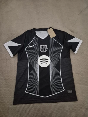Svart FC Barcelona tröja Nike - Snygg svart FC Barcelona matchtröja från Nike med vita grafiska linjer och klubbmärke på bröstet. Tröjan har korta ärmar, V-ringning och vit Spotify-logga framtill. Perfekt för fotbollsträning eller att bära som supporter.!Kan passa M!
