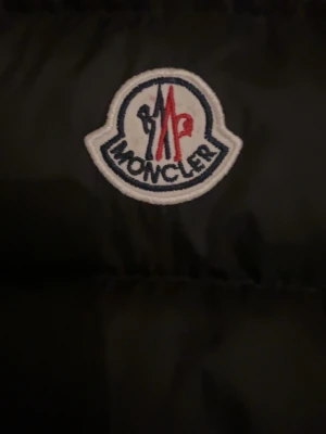 Svart dunväst från Moncler - Snygg svart dunväst från Moncler med klassisk logga på bröstet. Västen har hög krage, dragkedja framtill och två fickor med dragkedja. Perfekt för lager-på-lager och riktigt trendig look. Men en av fickorna med dragkedjor är sönder.