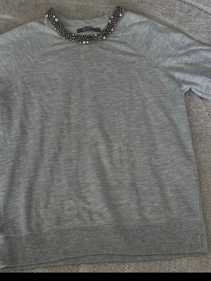 Grå sweatshirt med strass från Gina Tricot - Snygg grå sweatshirt från Gina Tricot med rund halsringning dekorerad med glittriga strassdetaljer. Tröjan har långa ärmar och ribbade muddar vid ärmslut och nederkant. Perfekt för dig som vill ha en basic tröja med lite extra bling.