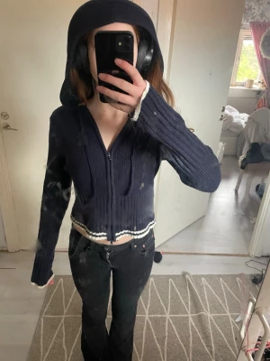 Mörkblå cropped hoodie med dragkedja - Säljer en mörkblå cropped hoodie med dragkedja framtill och ribbad struktur. Hoodien har vita detaljer vid ärmslut och nederkant samt en stor huva. Perfekt för en chill och avslappnad stil.