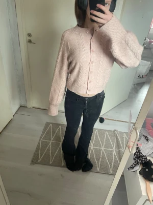Ljusrosa kabelstickad kofta - Söt ljusrosa kabelstickad kofta med rund hals och knappar framtill. Koftan har ballongärmar och ribbade muddar vid ärmslut och nederkant. Perfekt för en mysig och trendig look.