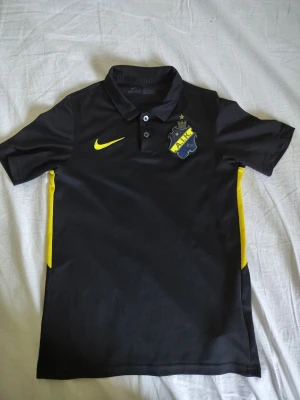 Svart AIK pikétröja från Nike - Svart pikétröja från Nike med AIK:s klubbmärke på bröstet och gul Nike-logga. Tröjan har krage med knappar och gula detaljer längs sidorna. Perfekt för dig som vill visa ditt AIK-stöd på träning eller match.