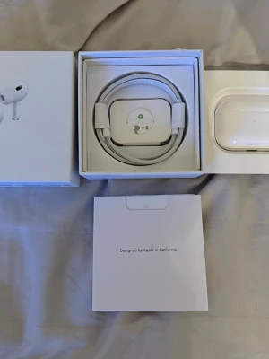 Apple AirPods Pro 2 med laddningsetui - Säljer ett par vita Apple AirPods Pro med trådlöst laddningsetui och tillhörande laddkabel. Hörlurarna har en stilren och kompakt design i blank plast och levereras i originalförpackning. Perfekta för musik, samtal och träning.