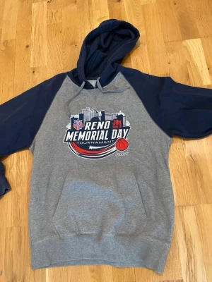 Grå och marinblå Reno Memorial hoodie stl S - Snygg hoodie med grå kropp och marinblå ärmar och huva. Stor tryckt grafik på bröstet med texten 'Reno Memorial Day Tournament 2019' och basketbollmotiv. Känguruficka framtill och dragsko i huvan. Perfekt för basketfans!