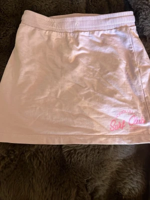 Ljusrosa kjol från Reaction - Söt ljusrosa kortkjol från Reaction med trycket 'Honolulu Surf Club' i rosa nertill. Kjolen har bred resår i midjan och är gjord i mjukt bomullsmaterial. Perfekt för en avslappnad och trendig look.
