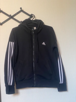 Svart Adidas hoodie med dragkedja - Svart hoodie från Adidas med vit logga på bröstet och klassiska vita ränder längs ärmarna. Tröjan har dragkedja framtill, två fickor och en huva med dragsko. Perfekt för en sportig och avslappnad stil.