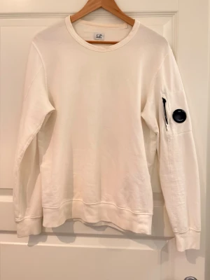 Vit sweatshirt från C.P. Company - Vit sweatshirt från C.P. Company med rund halsringning och ribbade muddar. Tröjan har en ikonisk svart linsdetalj och dragkedjeficka på ärmen. Perfekt för en clean och stilren look.