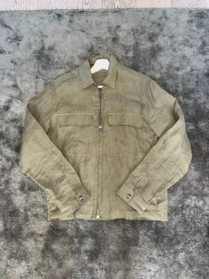 Beige overshirt från MNG i linne - Stilren beige overshirt från MNG i luftig linnemix. Jackan har två bröstfickor med lock, dragkedja framtill och klassisk krage. Perfekt för lager-på-lager och enkel att matcha med olika outfits.