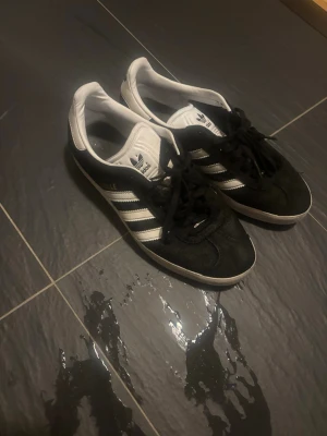 Adidas Gazelle svarta sneakers - Svarta Adidas Gazelle sneakers med klassiska vita ränder och vit sula. Ovandelen är i mocka och insidan är vit med Adidas-logga på plösen. Snygg och tidlös modell som passar till det mesta.