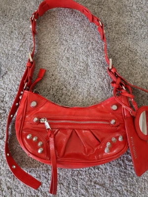 Röd Steve Madden handväska i skinn - Cool röd handväska från Steve Madden i glansigt skinn med silverfärgade nitar och dragkedja framtill. Justerbar axelrem med metalldetaljer och tillhörande spegel i matchande fodral. Perfekt statement piece för dig som gillar färg och detaljer.