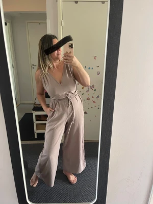 Beige jumpsuit med knytskärp - Snygg beige jumpsuit med djup V-ringning och vida ben. Jumpsuiten har ett matchande knytskärp i midjan och är ärmlös, vilket ger en avslappnad och trendig look. Tillverkad i ett mjukt och lätt tyg som känns skönt mot huden.Bomull.Helt ny med etikett 